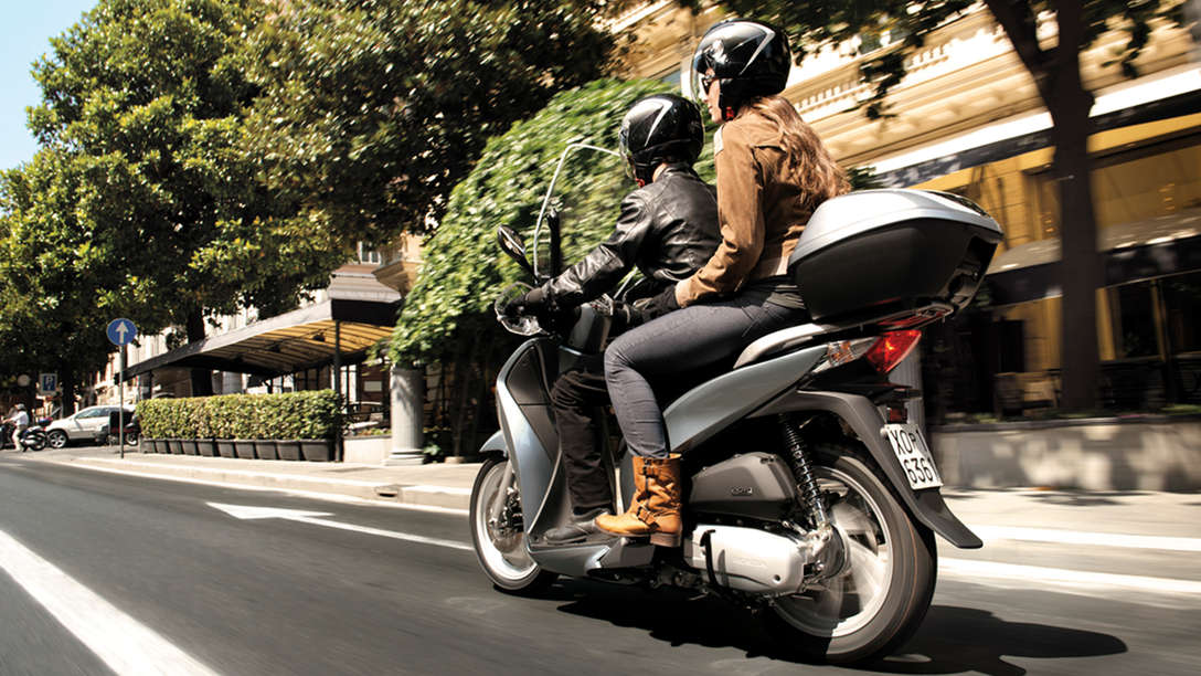 Honda SH125i - widok przedstawiający skuter z kierowcą i pasażerem z tylnej perspektywy. Skierowany w prawo (na drodze).