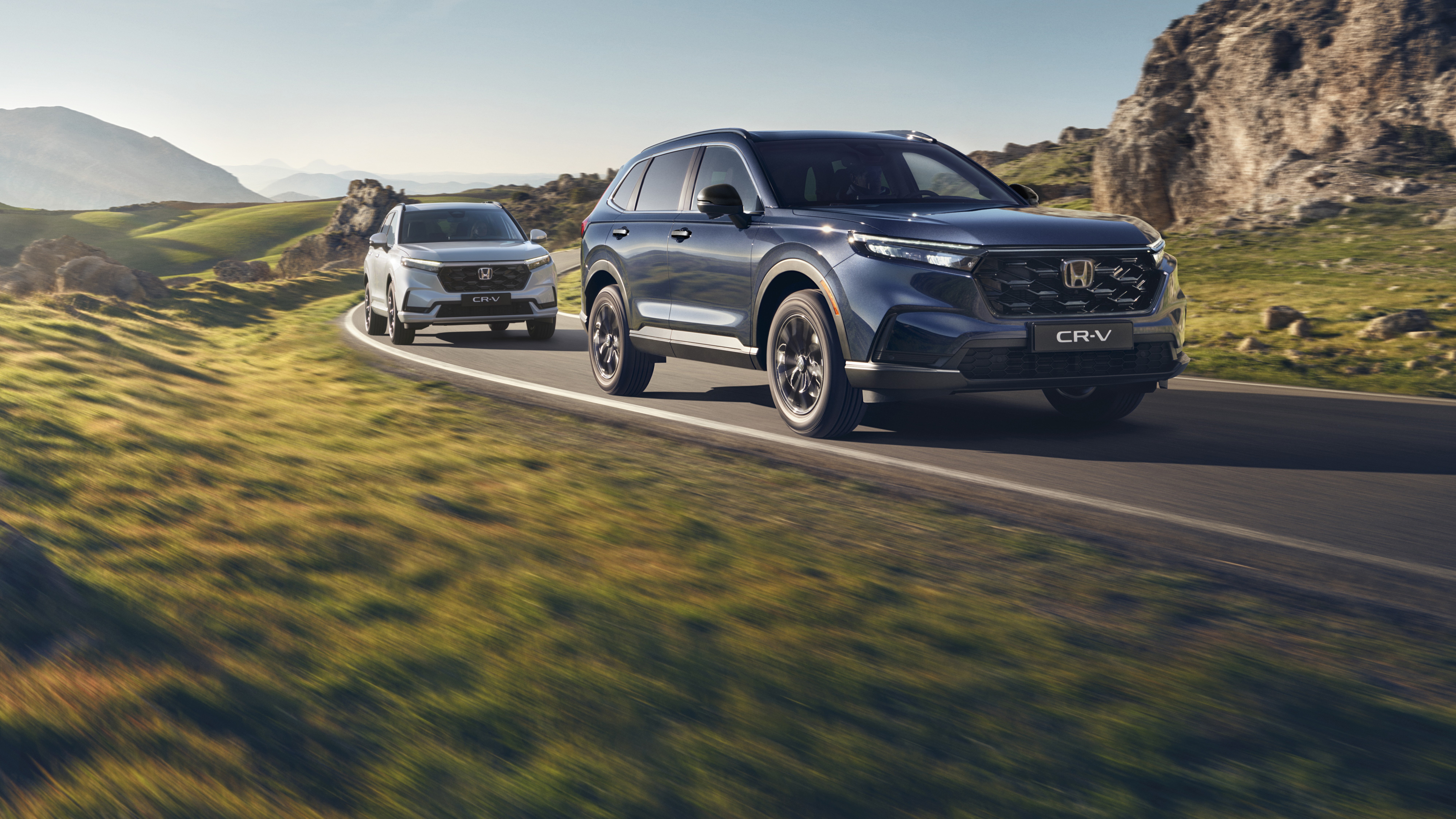 Srebrny SUV Honda CR-V jadący krętą górską drogą z skalistym terenem i górami w tle. Pojazd jest w ruchu, co stwarza poczucie prędkości i przygody w surowym krajobrazie.