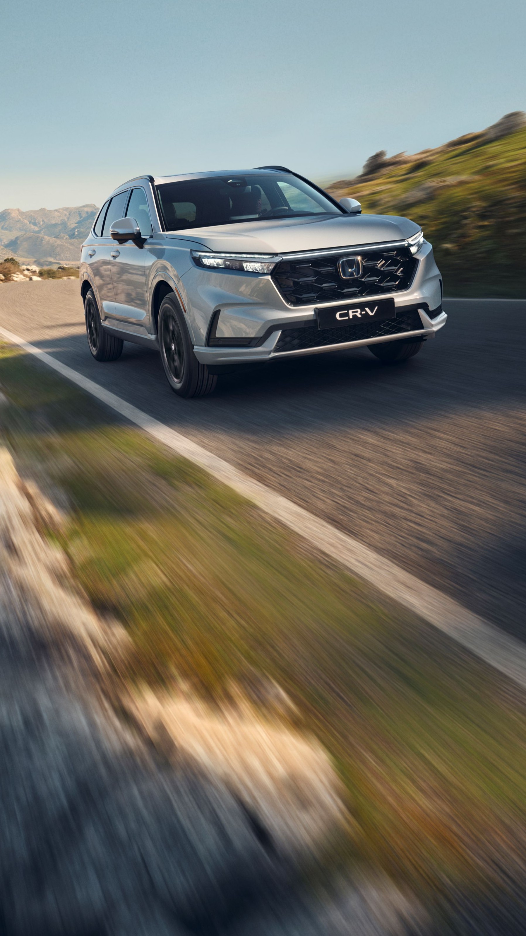 Srebrny SUV Honda CR-V jadący krętą górską drogą z skalistym terenem i górami w tle. Pojazd jest w ruchu, co stwarza poczucie prędkości i przygody w surowym krajobrazie.