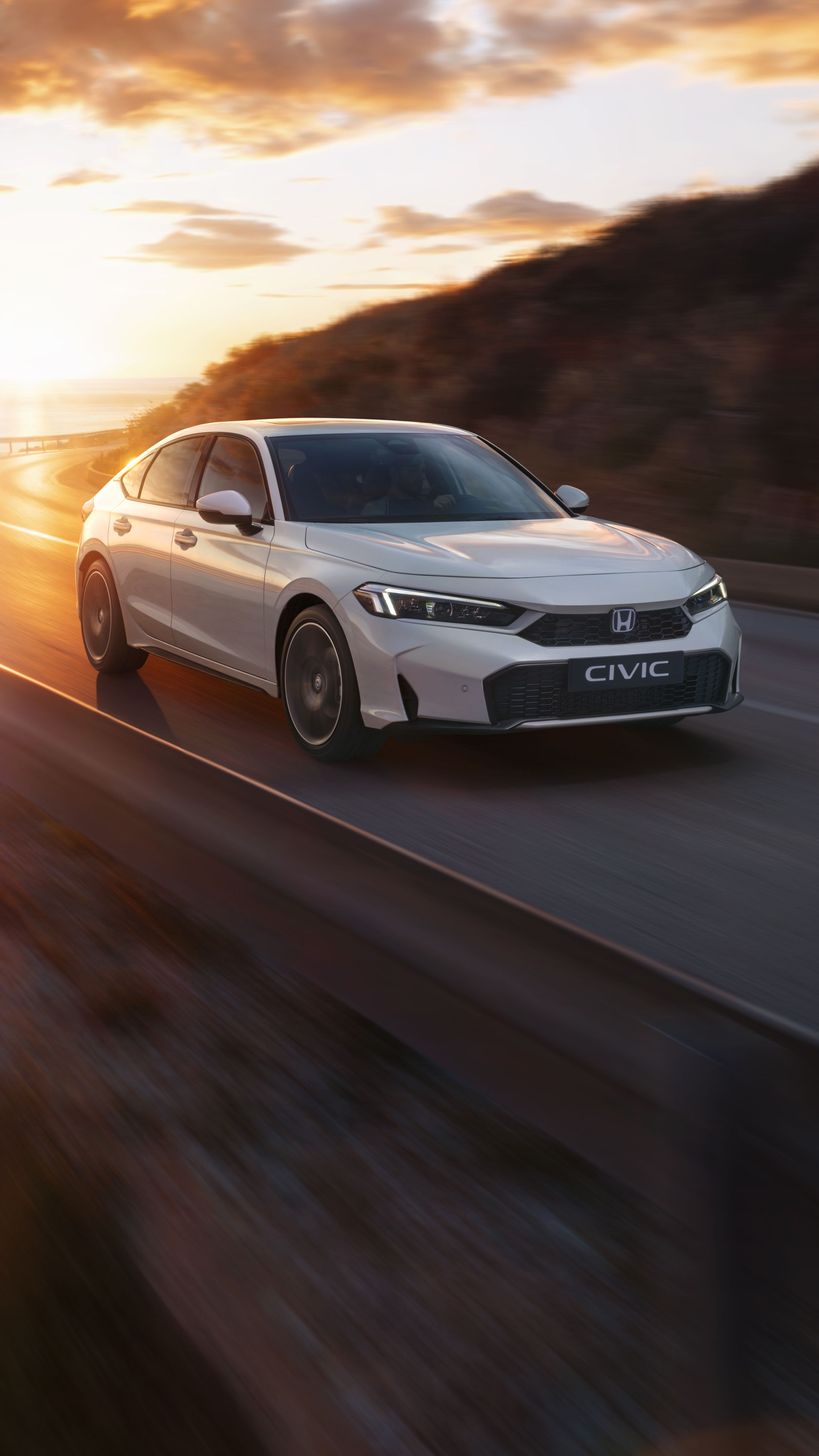 Biały Civic Hybrid jadący nadmorską drogą o zachodzie słońca, z morzem w tle.