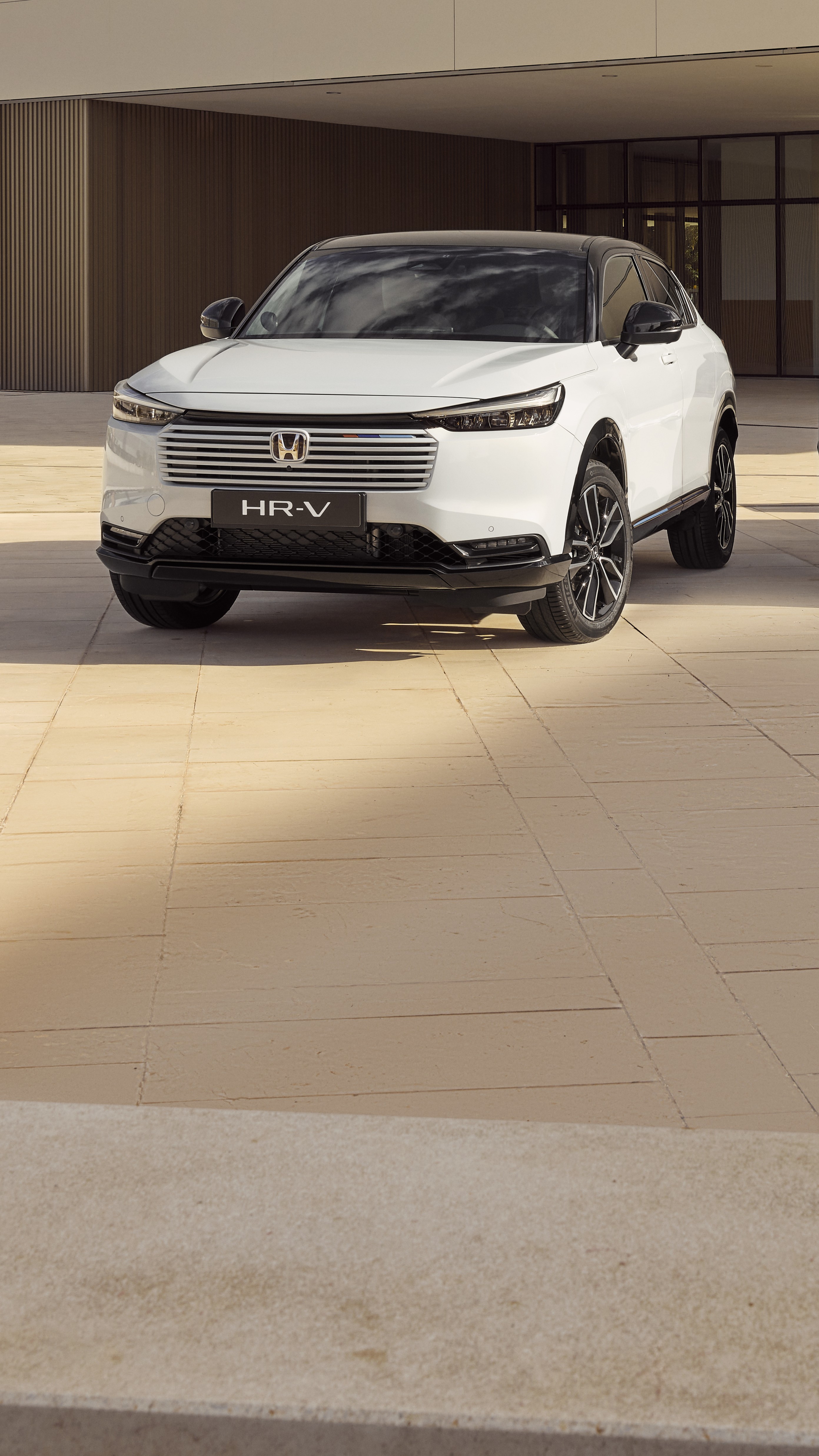 Biały SUV Honda HR-V zaparkowany przed nowoczesnym budynkiem z beżowymi kafelkowanymi ścianami. Pojazd jest ustawiony pod kątem, prezentując swój smukły wygląd i charakterystyczną osłonę chłodnicy.