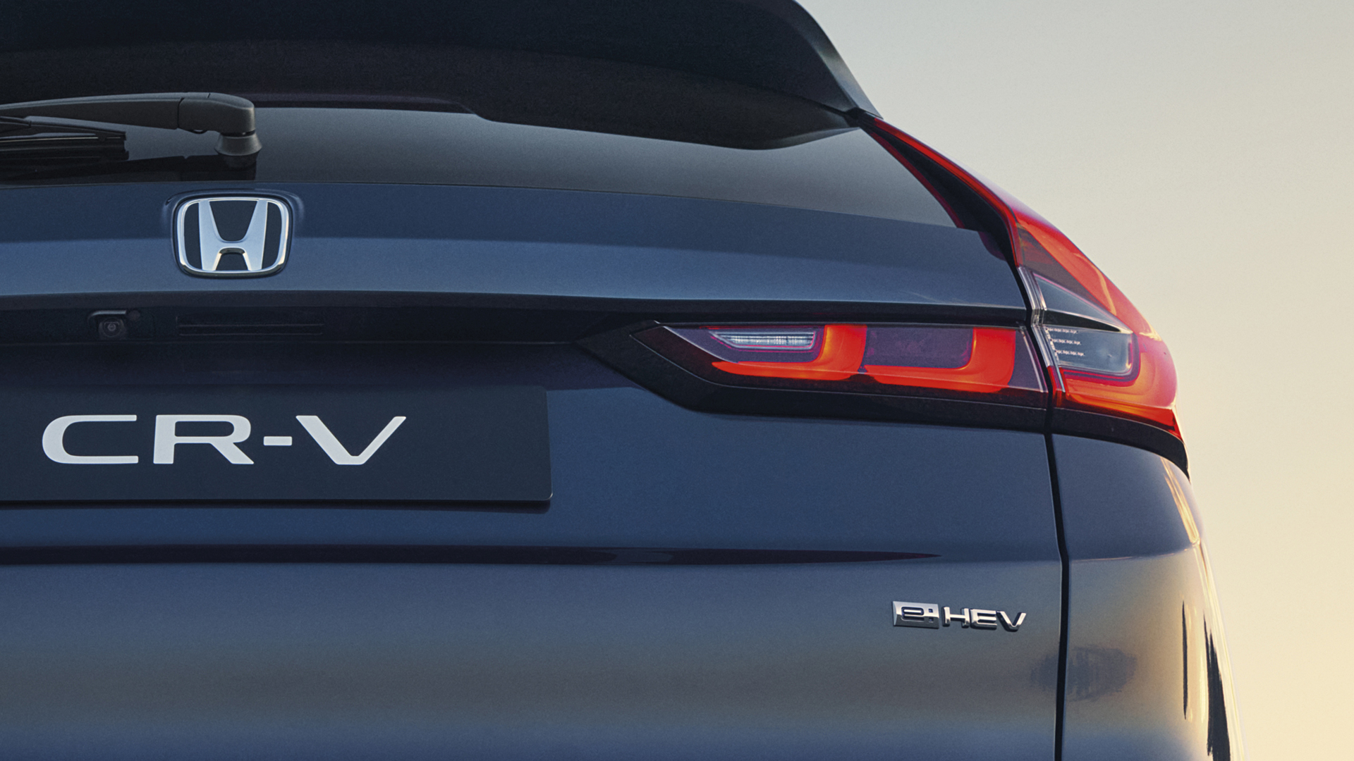 Zbliżenie na tylną część ciemnoniebieskiego Hondy CR-V e:HEV, ukazujące emblemat Hondy, oznaczenie CR-V oraz charakterystyczne, podświetlone LED-owe światła tylne na tle delikatnego nieba o zachodzie słońca.