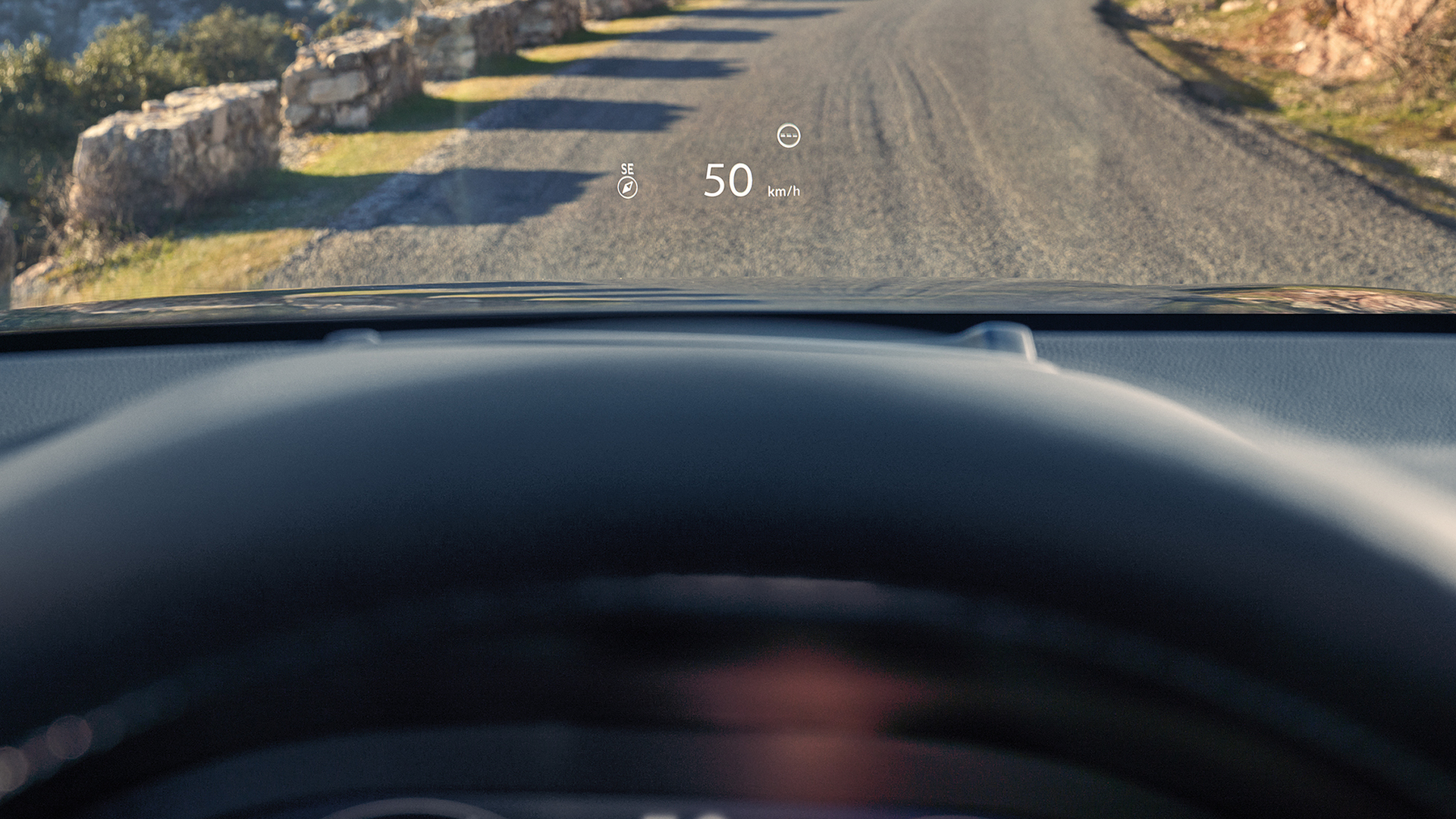 Widok z perspektywy pierwszej osoby z fotela kierowcy, przedstawiający wyświetlacz Head-Up Display (HUD), na którym widoczne są wskazania „50 km/h” oraz kompas wyświetlane na przedniej szybie nad żwirową drogą.