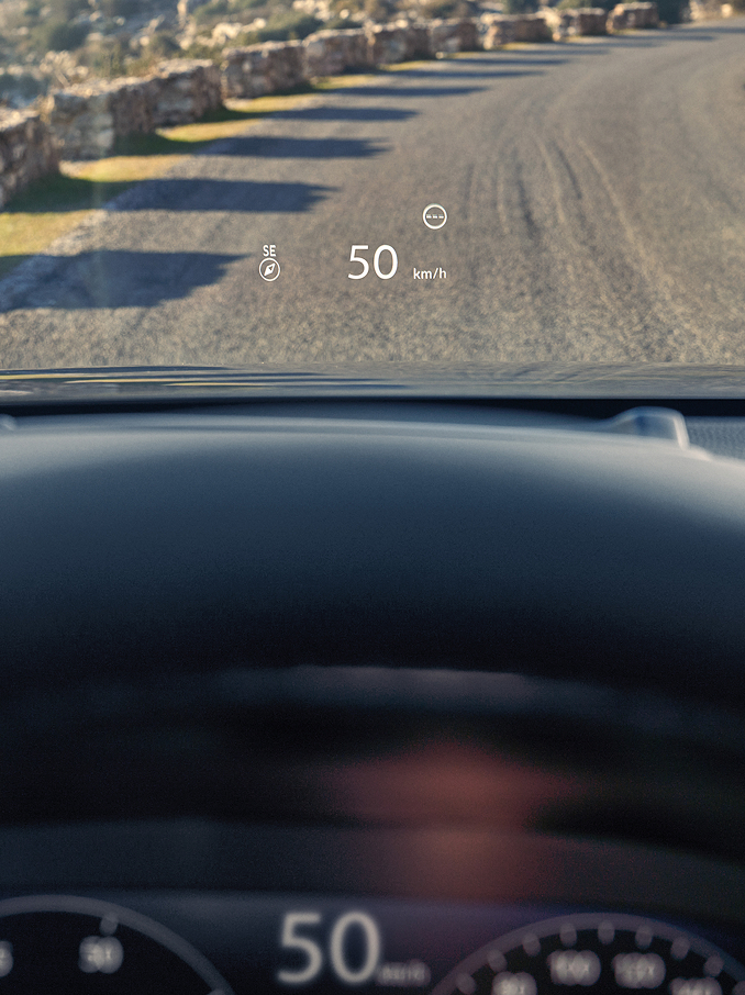 Widok z perspektywy pierwszej osoby z fotela kierowcy, przedstawiający wyświetlacz Head-Up Display (HUD), na którym widoczne są wskazania „50 km/h” oraz kompas wyświetlane na przedniej szybie nad żwirową drogą.
