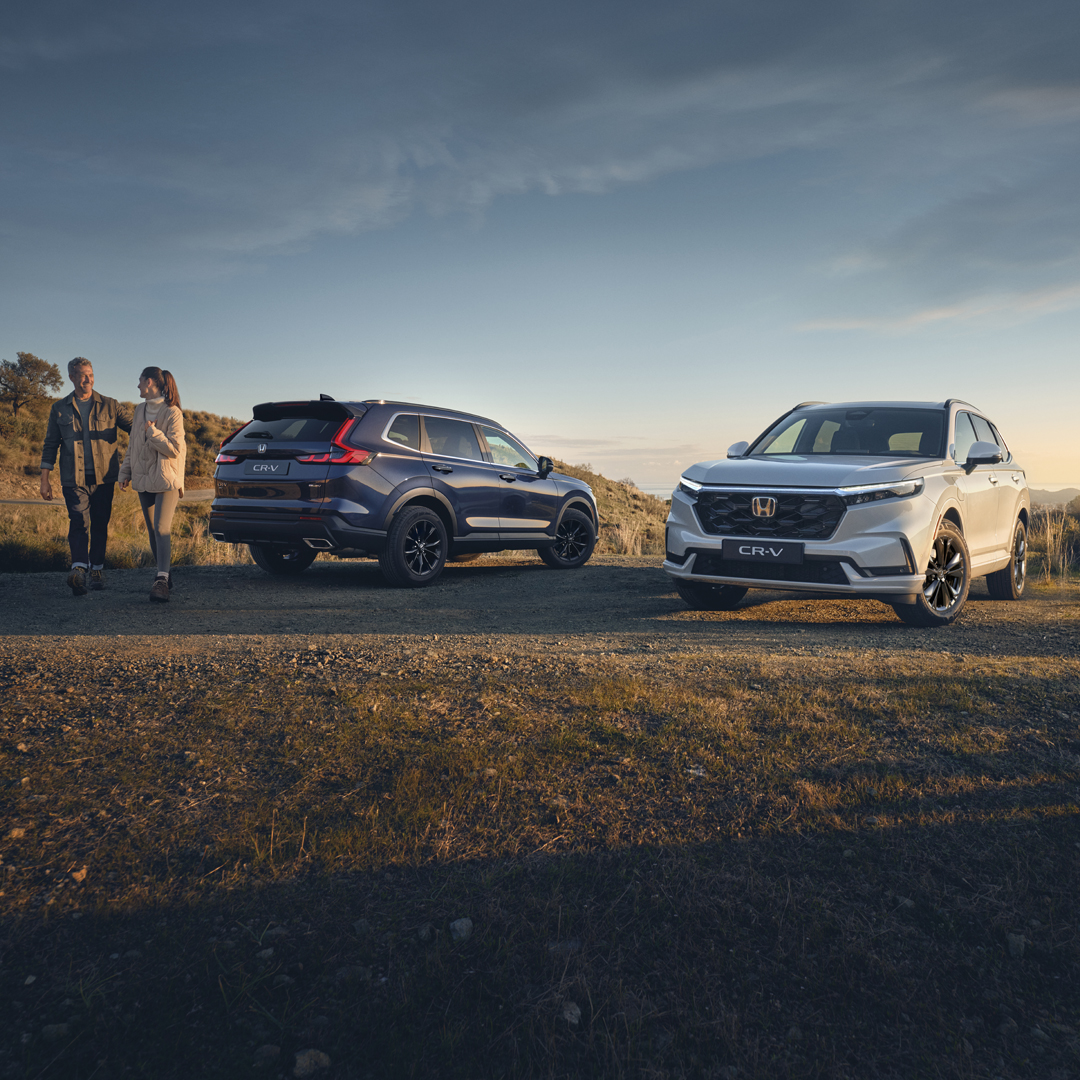 Dwa zaparkowane SUV-y — ciemnoniebieski i biały — stoją na polanie w wiejskim krajobrazie o złotej godzinie; w pobliżu spacerują dwie osoby, a w tle widać pagórki i wysoką trawę pod czystym niebem.