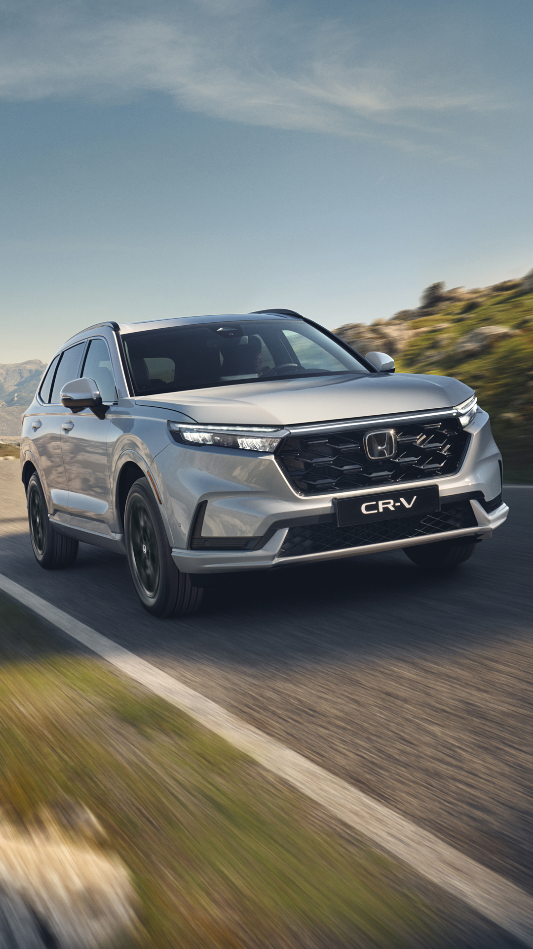 Srebrny SUV Honda CR-V jadący po krętej górskiej drodze wśród skalistego terenu, z górami w tle, pod czystym, błękitnym niebem.