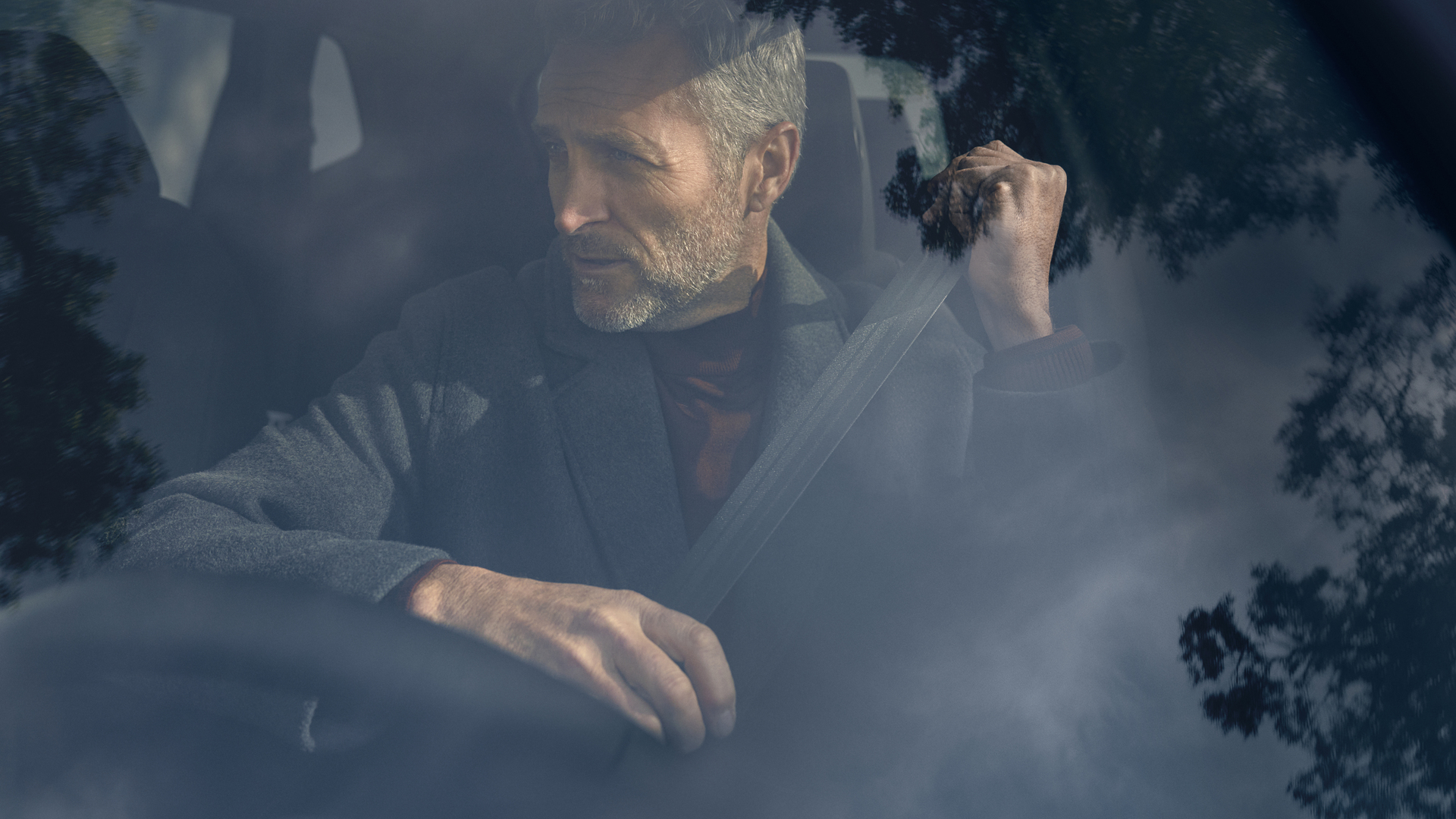 A gray-haired man sits in the driver's seat of a car, visible through a foggy or rain-covered windshield. He appears to be adjusting something near the rearview mirror with one hand while the other remains on the steering wheel.