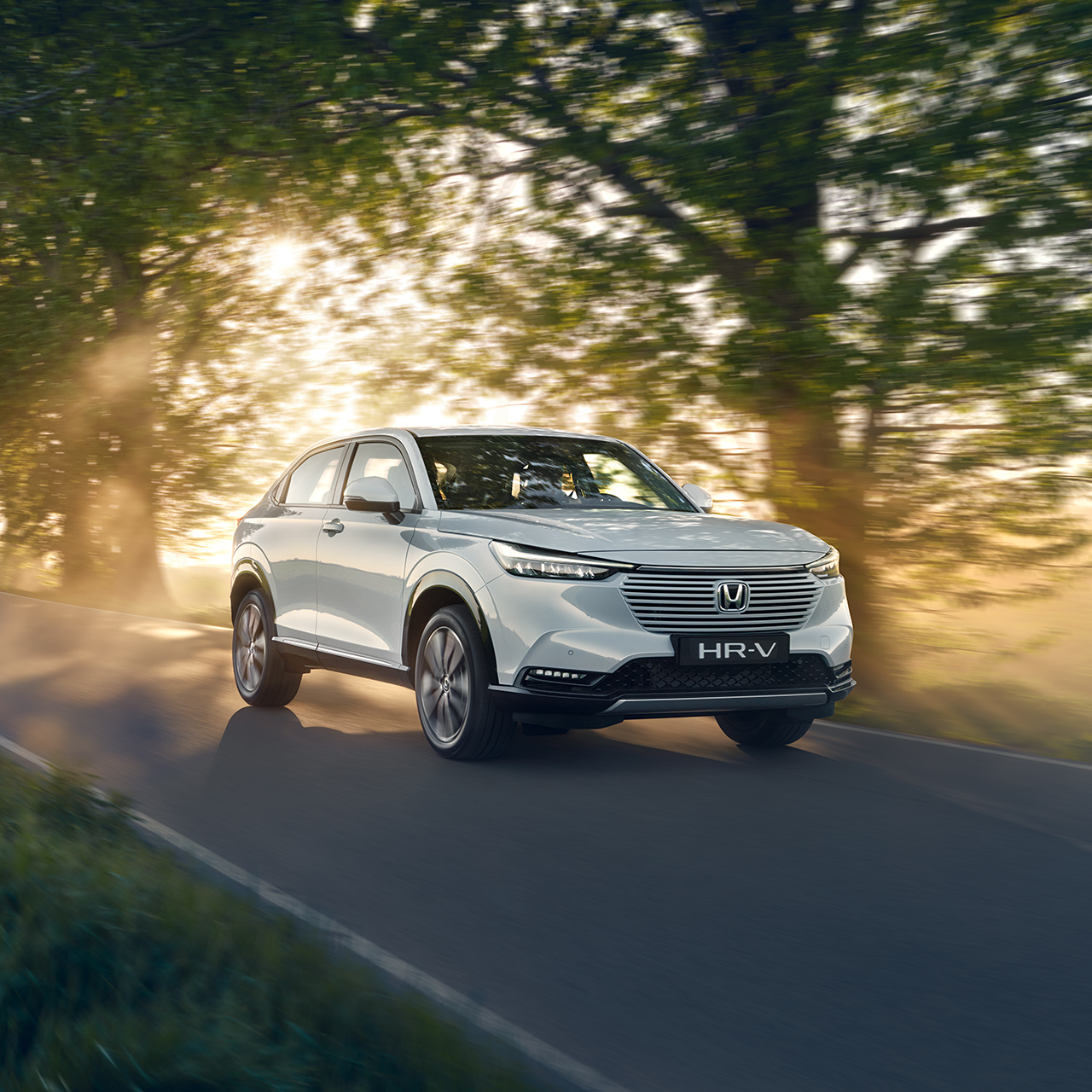 Hybrydowa Honda HR-V jadąca drogą w otoczeniu wiejskiego krajobrazu.