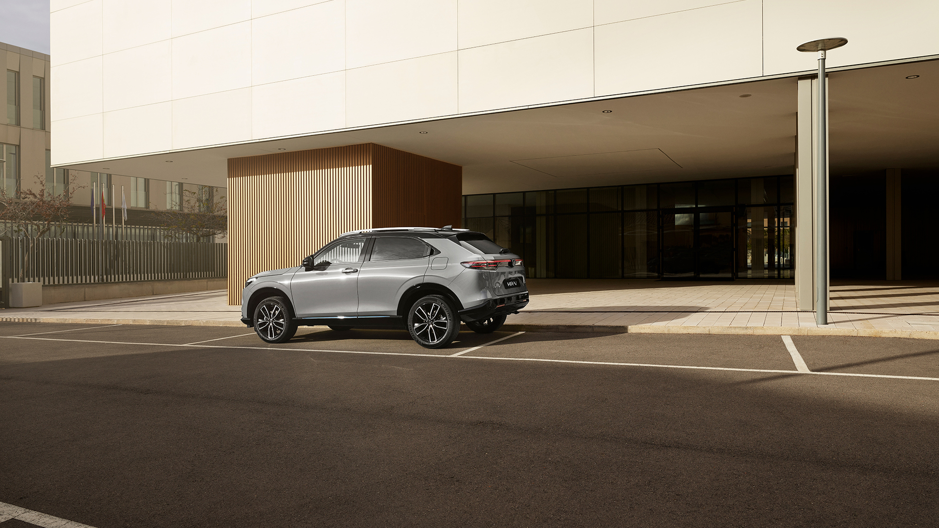 Srebrna Honda HR-V stoi na zewnętrznym parkingu przed nowoczesnym budynkiem ze szklanymi ścianami i pionowymi drewnianymi panelami, uchwycona od tyłu pod kątem trzech czwartych w świetle dziennym.