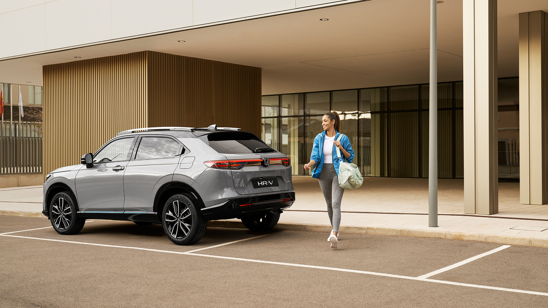 Srebrny Honda HR-V zaparkowany przed nowoczesnym budynkiem, obok którego przechodzi kobieta z torbą – scena podkreślająca wygodę i praktyczność auta w codziennym, miejskim życiu.