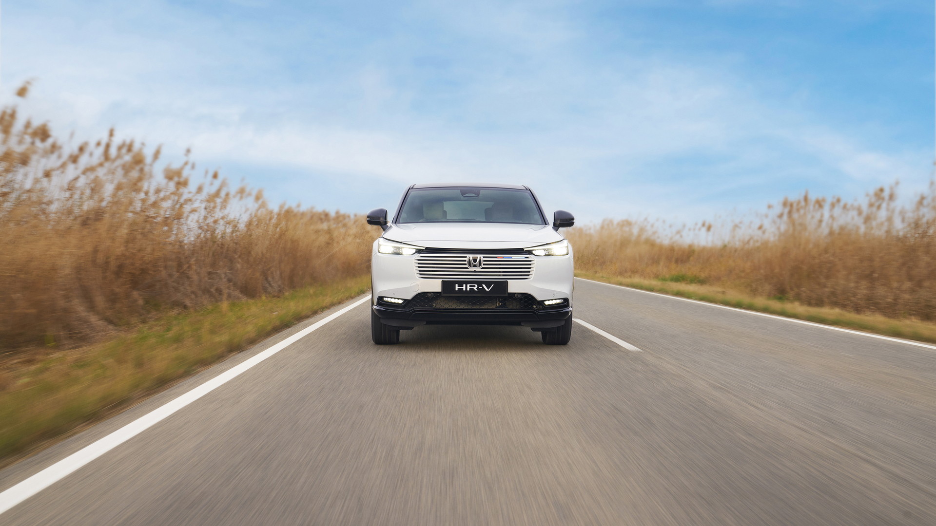 Biały Honda HR-V jedzie w kierunku kamery po dwupasmowej drodze, otoczonej wysoką, złotą trawą, pod błękitnym niebem.