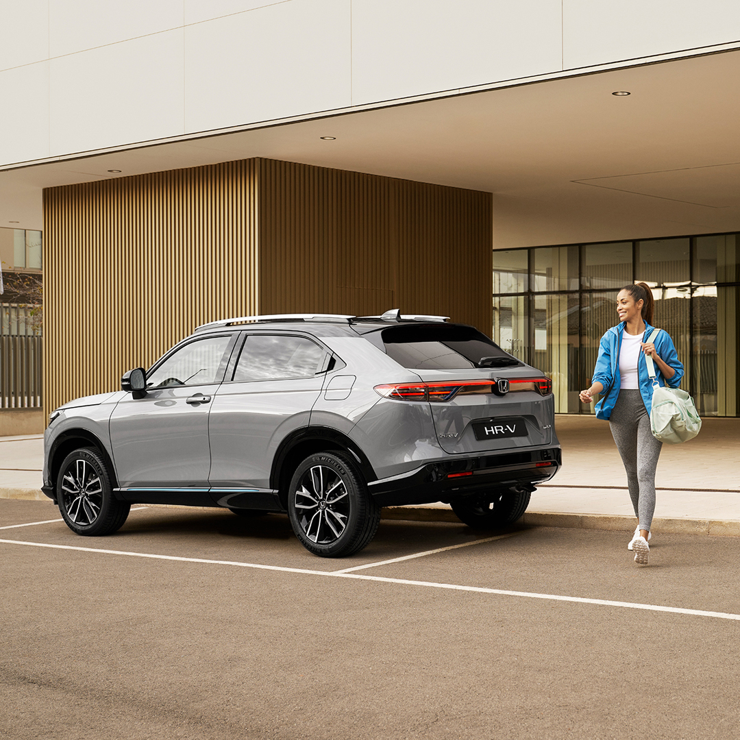 Srebrny Honda HR-V zaparkowany przed nowoczesnym budynkiem, obok którego przechodzi kobieta z torbą – scena podkreślająca wygodę i praktyczność auta w codziennym, miejskim życiu.