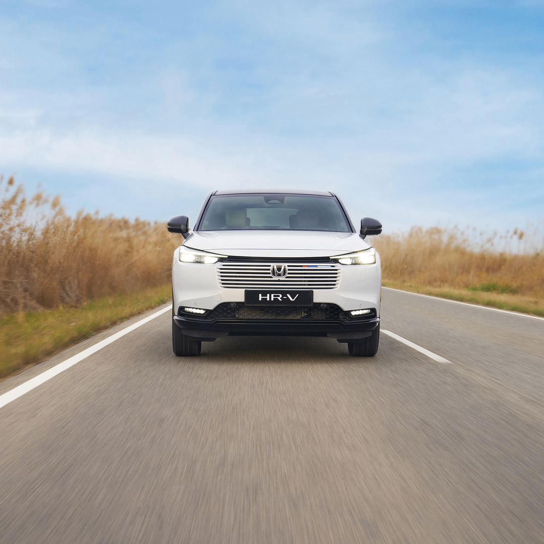 Biały Honda HR-V jedzie w kierunku kamery po dwupasmowej drodze, otoczonej wysoką, złotą trawą, pod błękitnym niebem.