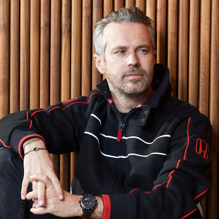 Mężczyzna w czapce i kurtce Honda Racing, dotykający ronda czapki.