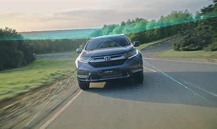 Honda Sensing — samochód w podmiejskiej okolicy z ilustracją systemu zapobiegania zjeżdżaniu z drogi oraz rozpoznawania znaków drogowych.