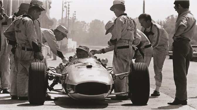 Bolid Formuły 1 z lat 60. wraz z kierowcą i inżynierami, widok z przodu.