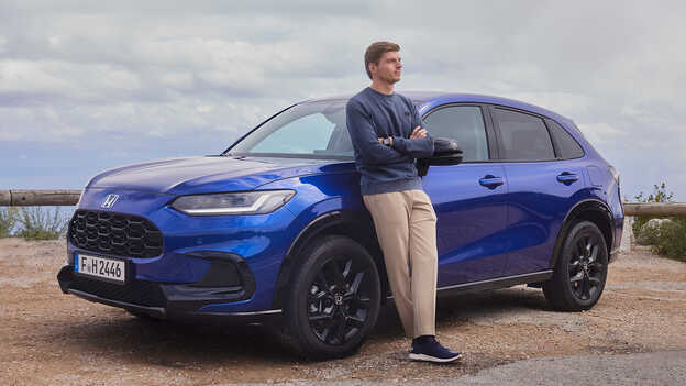 Max Verstappen opierający się o zaparkowanego niebieskiego hybrydowego SUVa Honda ZR-V eksponującego swoją sportową estetykę.