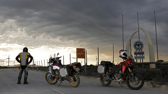 Kierowca patrzy się w horyzont, stojąc obok dwóch motocykli Africa Twin zaparkowanych na asfalcie.