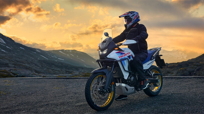 Model jadący motocyklem XL750 Transalp w górskiej scenerii.