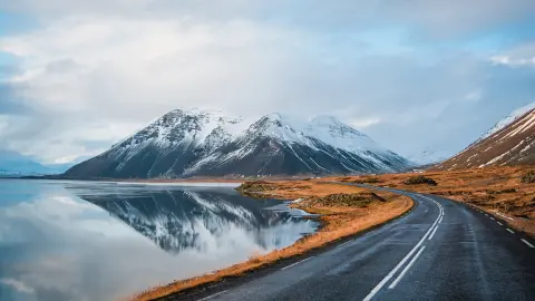 Panoramiczne zimowe zdjęcie drogi prowadzącej nad brzegiem jeziora do wulkanicznych gór. Wysokie skaliste szczyty pokryte warstwą śniegu odbijające się na tafli wody. Widok zza kierownicy na Obwodnicę, Islandia.