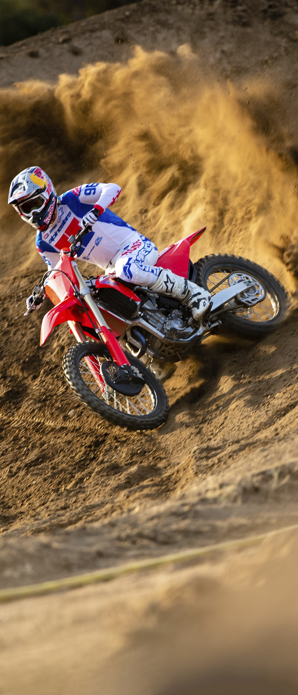 Kierowca motocrossu w biało-niebiesko-czerwonym stroju wzbija w powietrze falę piachu, wjeżdżając czerwonym motocyklem terenowym w stromy, piaszczysty zakręt.