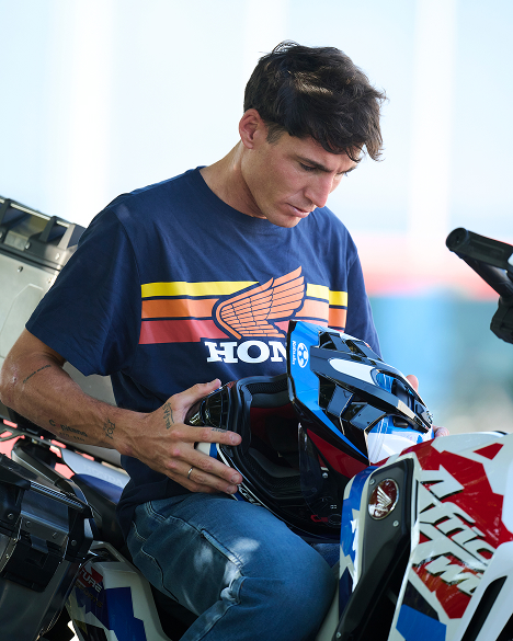 A person wearing a blue Honda t-shirt with retro-style stripes and logo examines a blue and white Honda motorcycle, likely a sportbike model. The individual is leaning over the bike, focusing intently on some aspect of it.