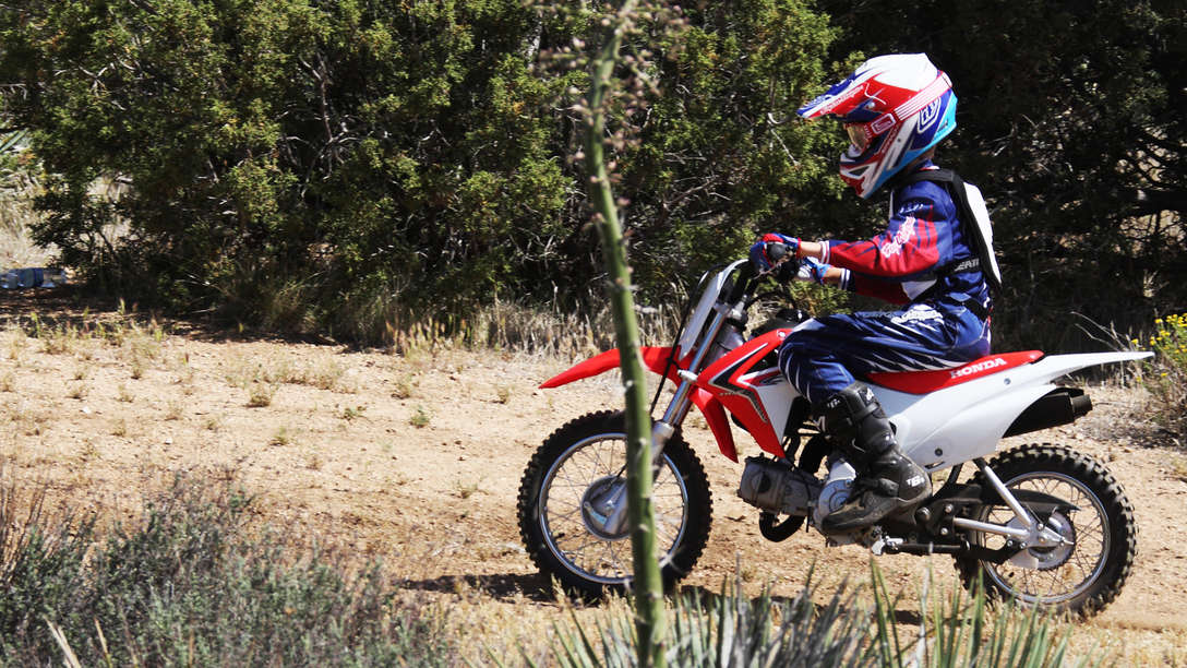 CRF110F — widok z boku 3/4 z kierowcą na torze motocrossowym. Skierowany w lewo.