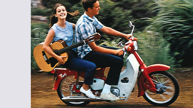 Honda Cub z młodym pasażerem wiozącym gitarę. Zdjęcie na drodze polnej. Motocykl skierowany w prawo.